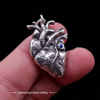 Sterling silver anatomical heart pendant by RBZ Jewelry featuring a custom natural blue sapphire gemstone setting, held between fingers for scale to showcase the realistic biological textures and high-contrast oxidized finish.