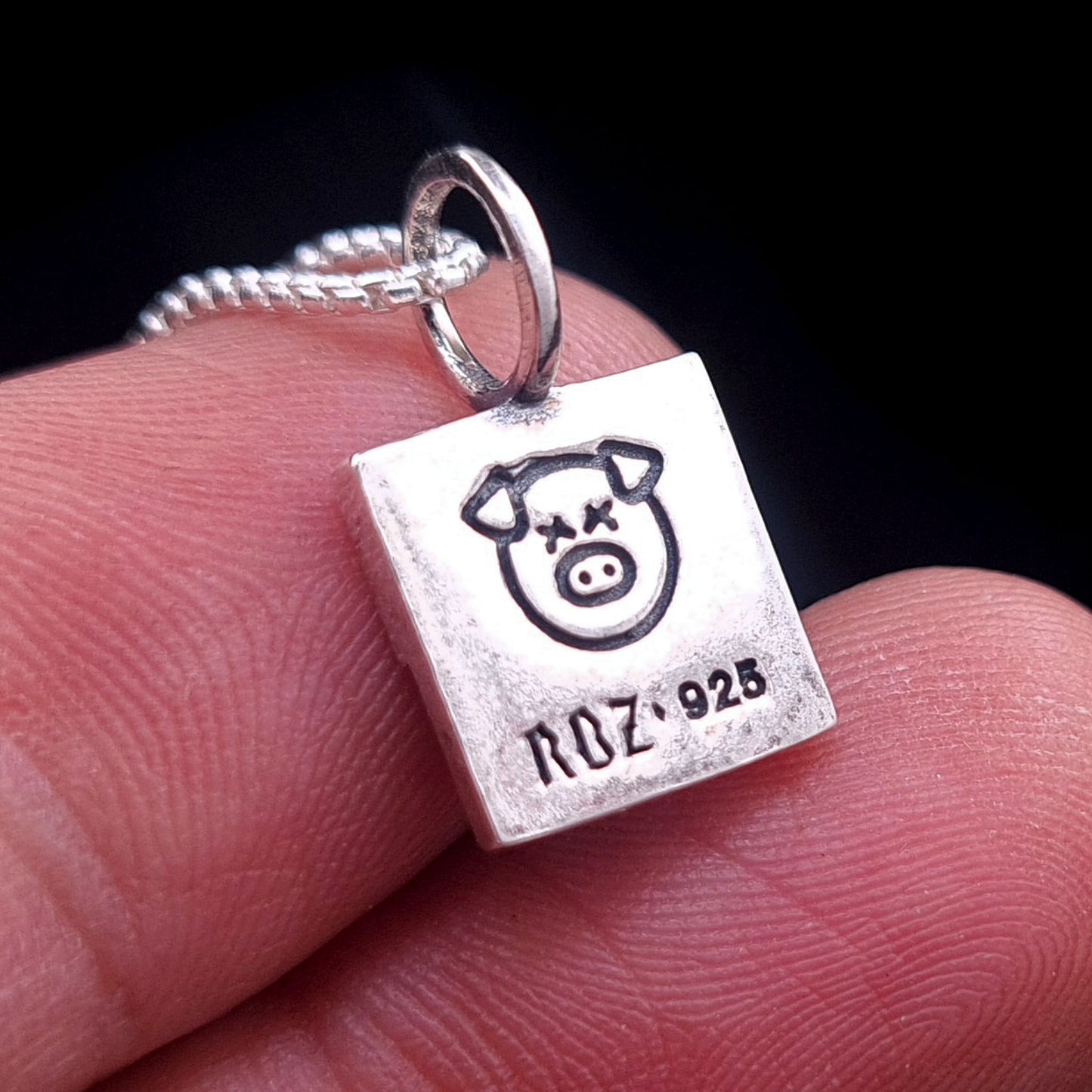 Close-up of the reverse side of the sterling silver ACAB pendant held in hand, showing the engraved X-eyed pig graphic and RBZ 925 hallmark on a brushed brutalist metal finish.