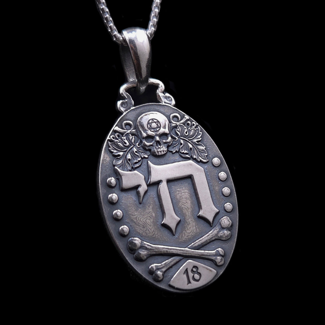 Gothic Judaica sterling silver Chai pendant by RBZ Jewelry, featuring high-relief Hebrew letters חי, a detailed skull engraved with a Star of David, vine leaves, crossed bones, and the number 18 plaque on a deeply oxidized oval medallion.