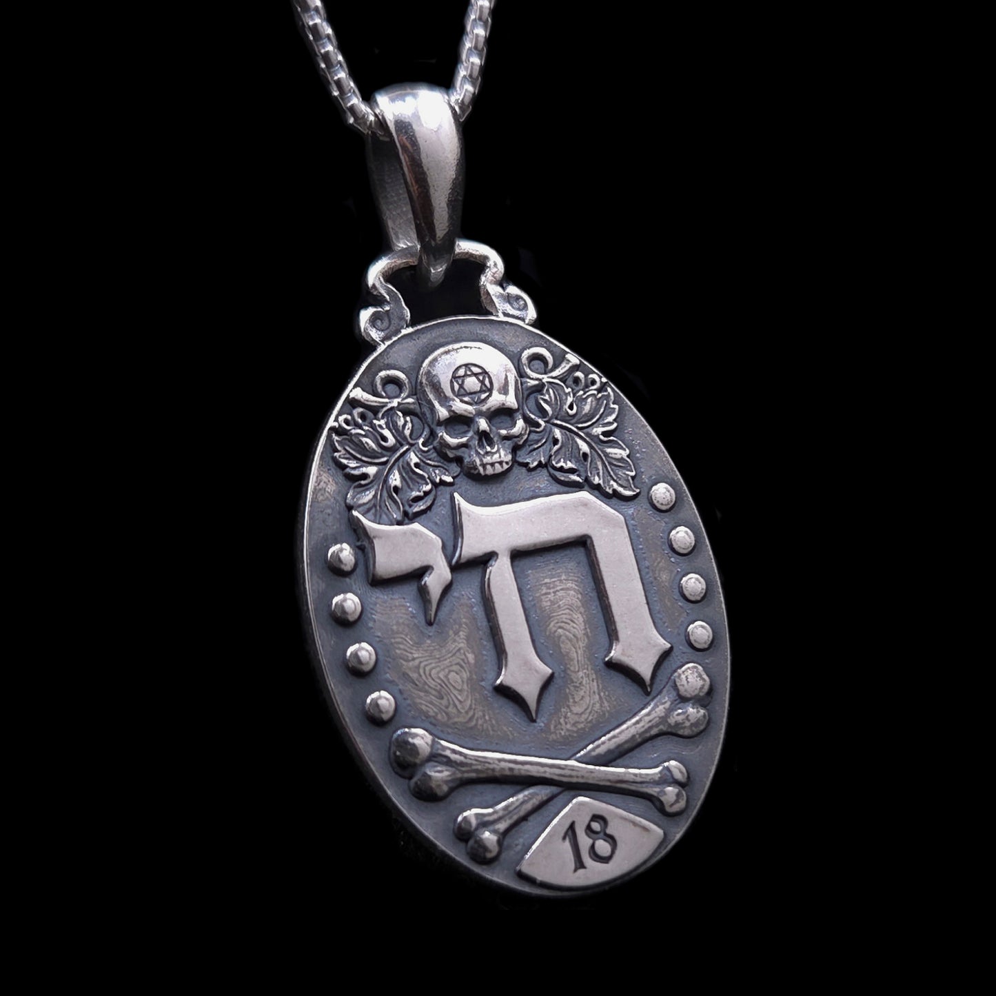Gothic Judaica sterling silver Chai pendant by RBZ Jewelry, featuring high-relief Hebrew letters חי, a detailed skull engraved with a Star of David, vine leaves, crossed bones, and the number 18 plaque on a deeply oxidized oval medallion.