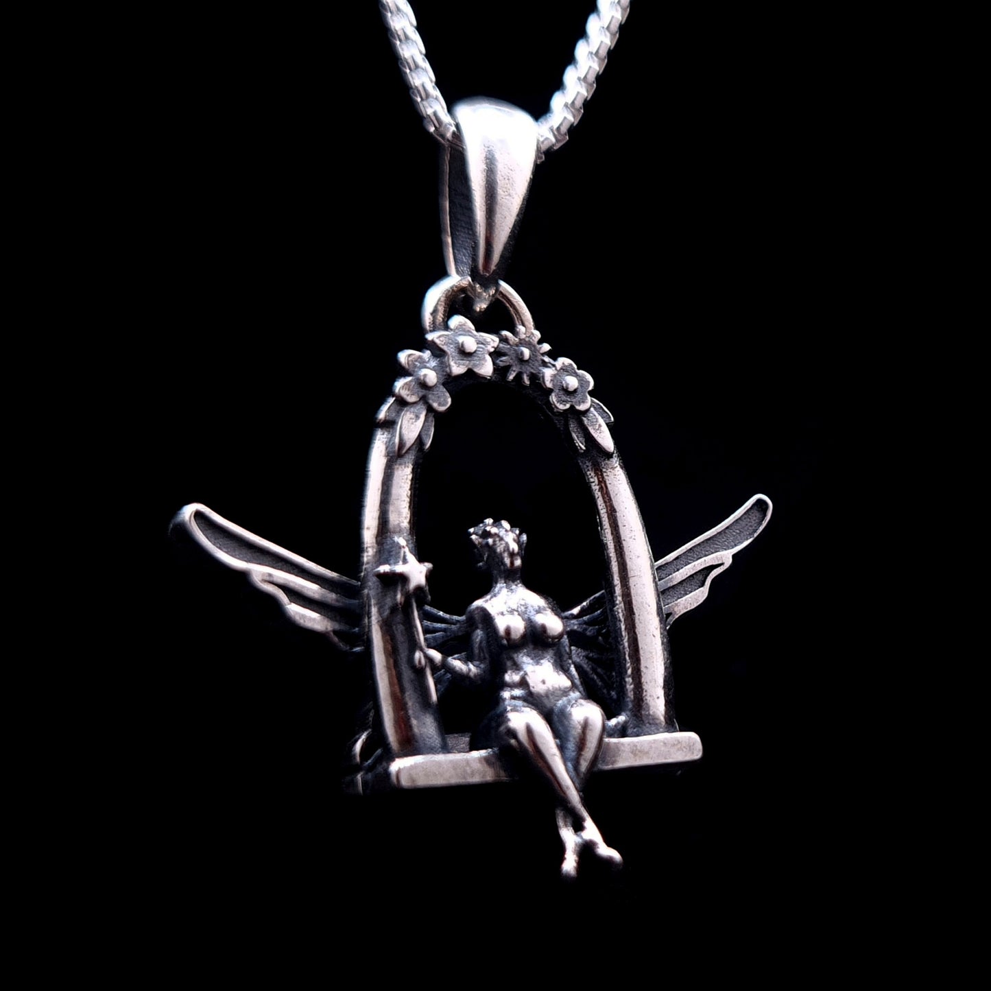 Sterling silver Flower Fairy pendant by RBZ Jewelry, featuring an intricately detailed fairy with spread wings sitting on a floral-arched swing and holding a star-tipped wand against a black background.