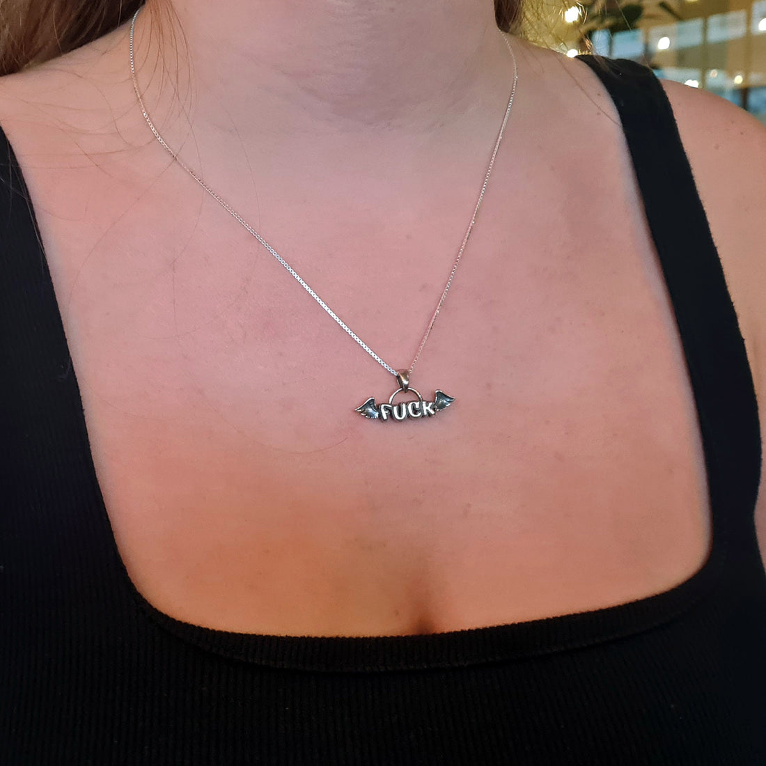 A female model wearing the sterling silver Flying Fuck pendant by RBZ Jewelry on a silver chain, styled with a black square-neck top to demonstrate the necklace length and scale.