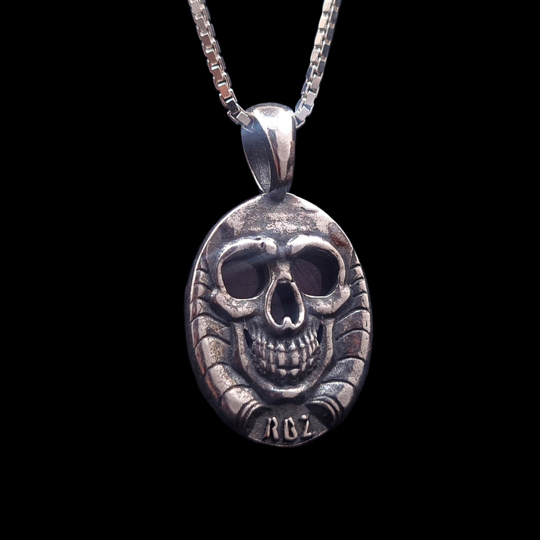 Studio-crafted RBZ Jewelry Gothic pendant back view, featuring a detailed Sterling silver grinning skull with hollow eyes and nose revealing the natural African Ruby's glow, part of the double-sided Skull Bezel design, with the 'RBZ' logo hallmark clearly visible.