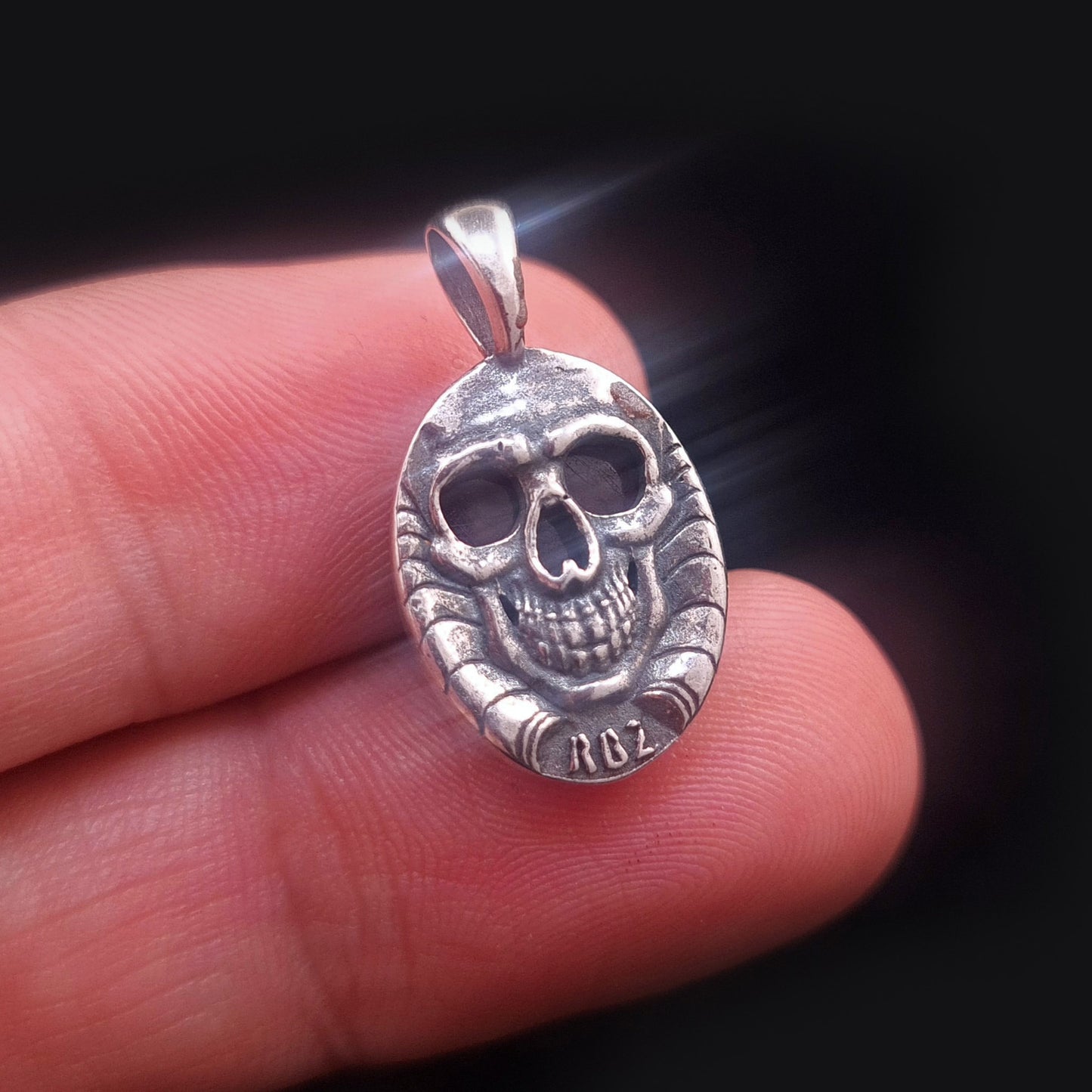 Studio-crafted RBZ Jewelry Gothic pendant reverse view, held between fingers for scale, showcasing the detailed Sterling silver grinning skull. The deep crimson glow of the Natural African Ruby is visible through the skull's hollow eye sockets and nasal cavity, confirming its function as the reverse of the double-sided Skull Bezel design.