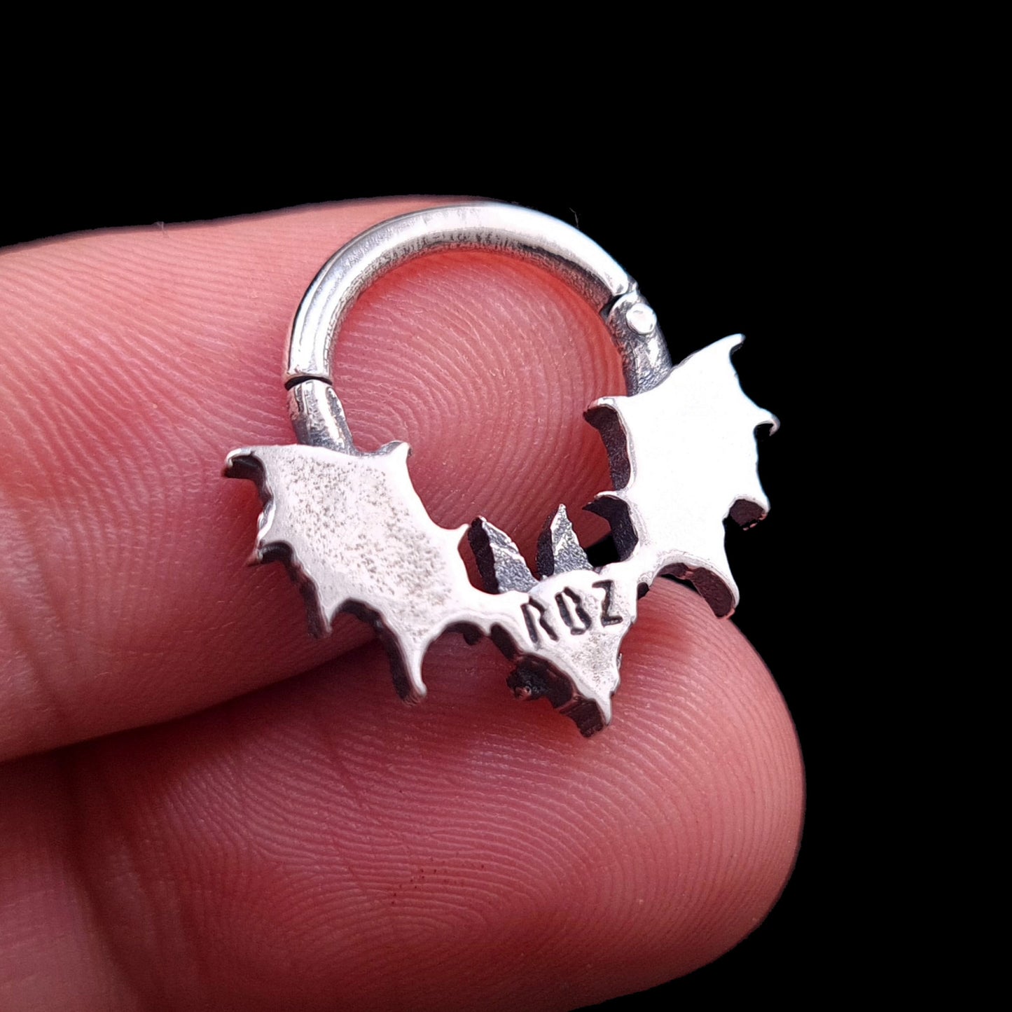 A close-up, reverse view of the Gothic Sterling silver Glitched Bat clicker septum ring by RBZ Jewelry, held between the fingers of a hand. The polished back of the distorted bat silhouette prominently features a clear "RBZ" hallmark stamp. The ring is closed against a black background.