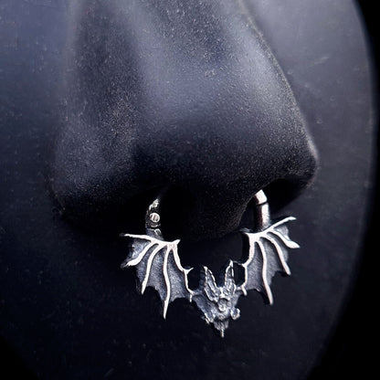A studio-crafted RBZ Jewelry Gothic Sterling silver Glitched Bat clicker septum ring shown worn on a life-size black silicone nose to demonstrate fit and appearance. The piece features a visceral, distorted bat silhouette with textured wings.