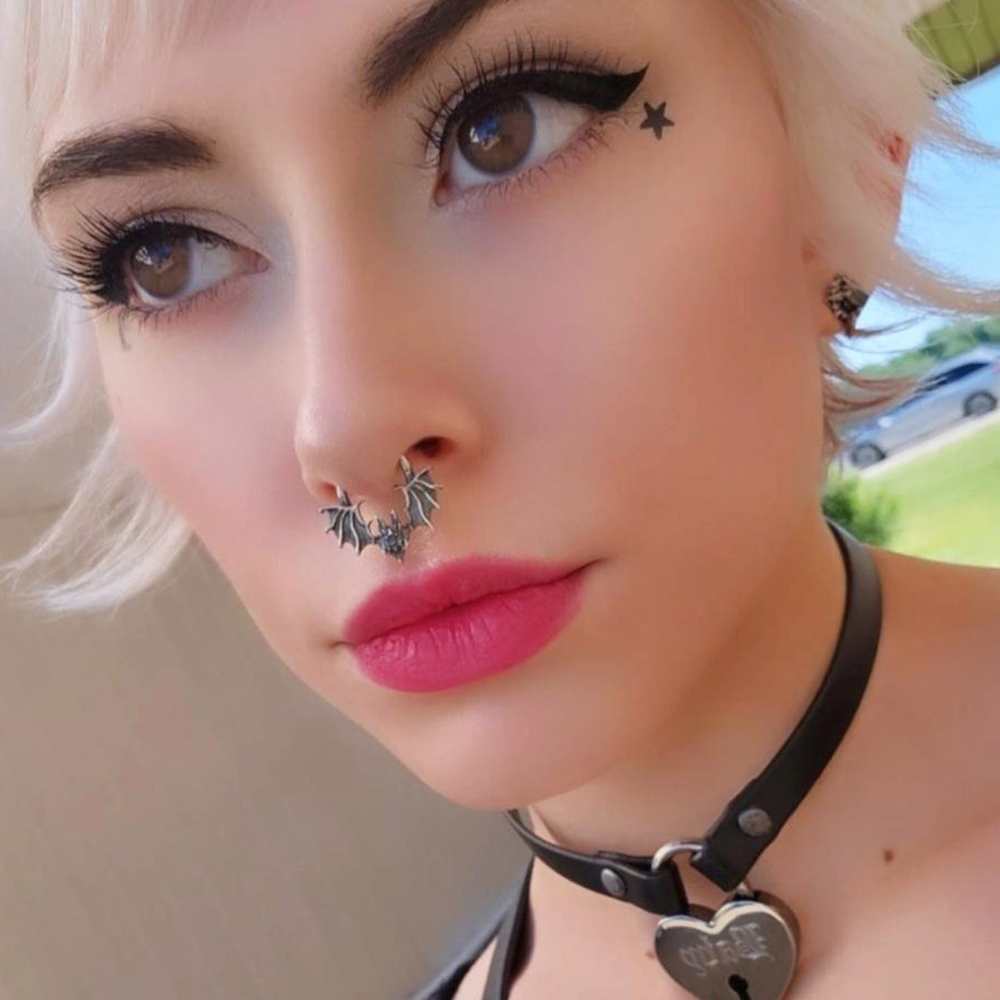 Close-up portrait of a female model with short blonde hair and bold cat-eye make-up, wearing the visceral Gothic Sterling silver Glitched Bat clicker septum ring by RBZ Jewelry. The intricate, distorted bat silhouette sits centrally, complemented by a star cheek tattoo and a black heart-lock choker.