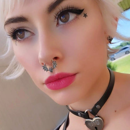 Close-up portrait of a female model with short blonde hair and bold cat-eye make-up, wearing the visceral Gothic Sterling silver Glitched Bat clicker septum ring by RBZ Jewelry. The intricate, distorted bat silhouette sits centrally, complemented by a star cheek tattoo and a black heart-lock choker.