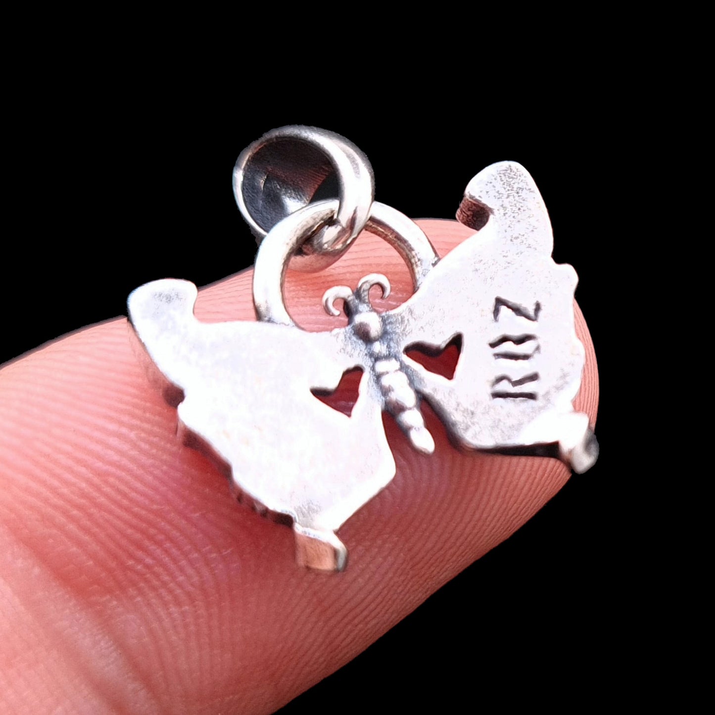 Rear view of the RBZ Jewelry Sterling silver Gothic Butterfly Skull pendant held on a finger for scale, showing the engraved logo and heart cutouts against a black background.