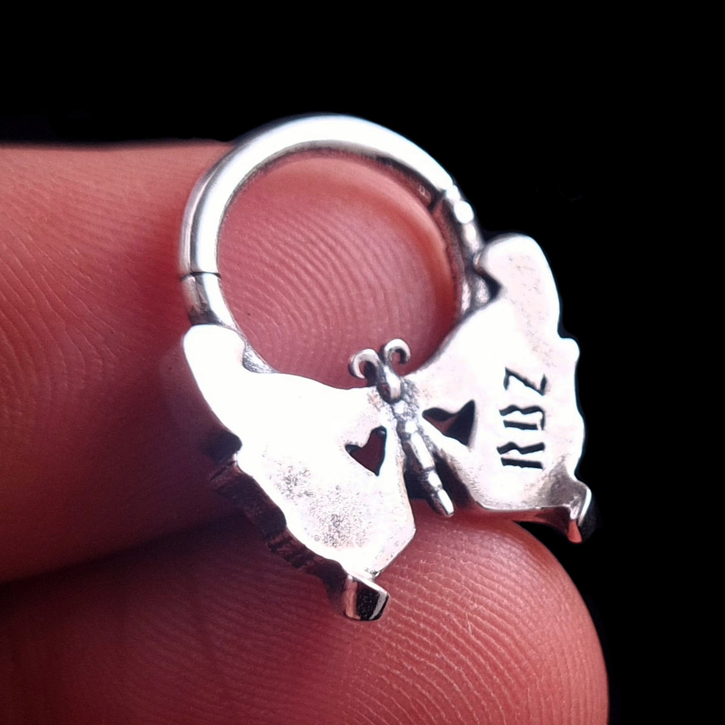 Detailed back view of the Sterling silver Gothic butterfly skull septum clicker ring by RBZ Jewelry, held between fingers to show scale, featuring the polished smooth reverse side of the wings with an visible engraved 'RBZ' logo.