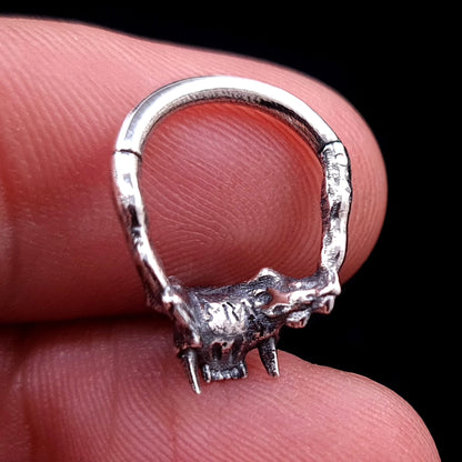 Macro back view of the RBZ Jewelry sterling silver Cat Skull septum ring, held between a finger and thumb, detailing the textured anatomical skull structure, fangs, clicker hinge mechanism, and the 'RBZ' logo engraving from behind, with visible fingerprints for scale.
