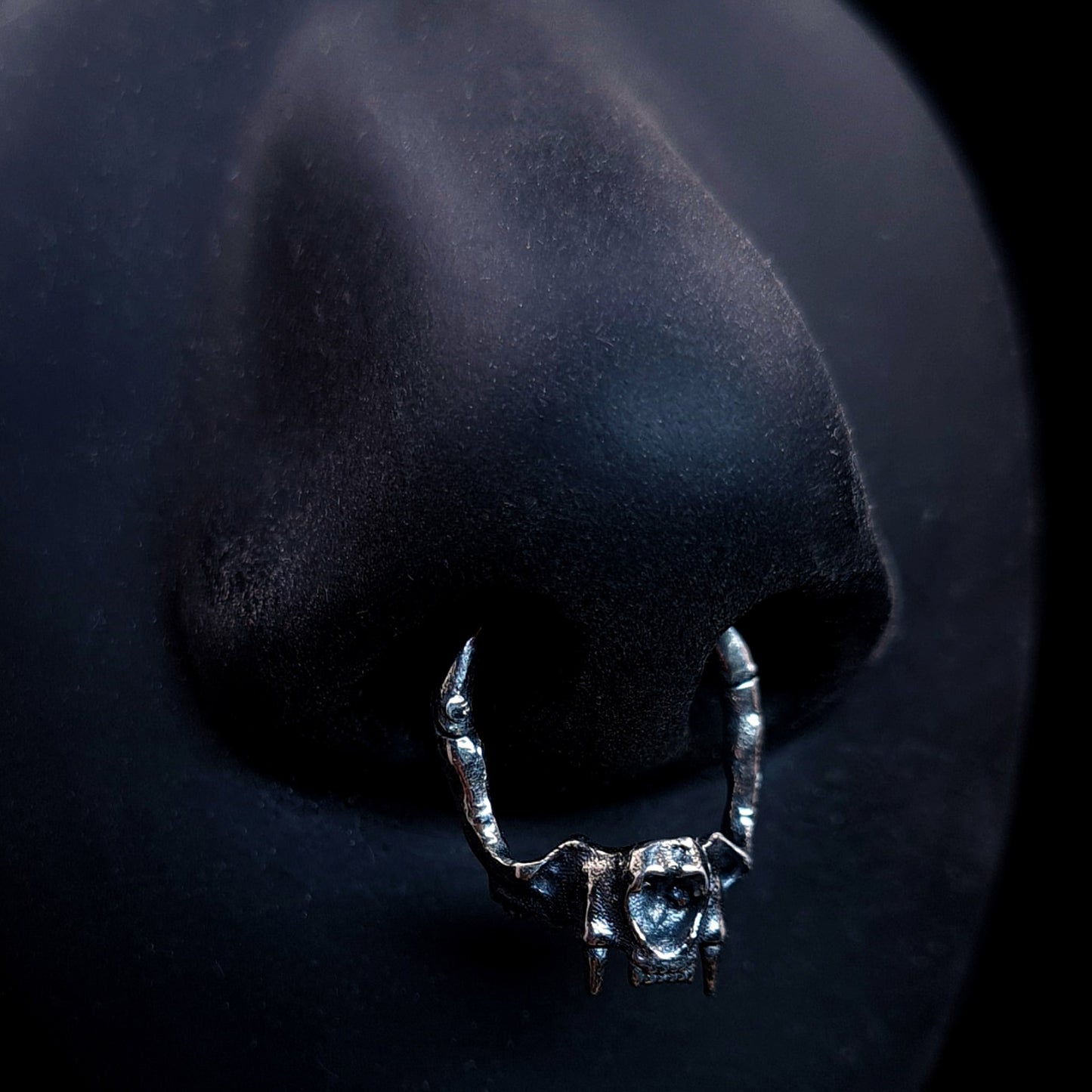 Product shot of the RBZ Jewelry sterling silver cat skull septum clicker ring worn on a life-size matte black silicone nose, demonstrating the fit and anatomical bone texture of the gothic predator design.