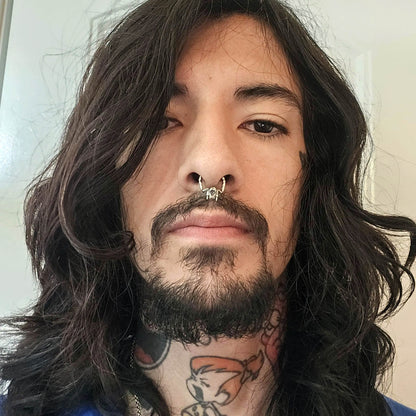 Front portrait of a tattooed man with long dark hair and a beard, wearing the RBZ Jewelry sterling silver Cat Skull septum clicker ring. The detailed cat skull with fangs and bone texture is prominently displayed in his septum.