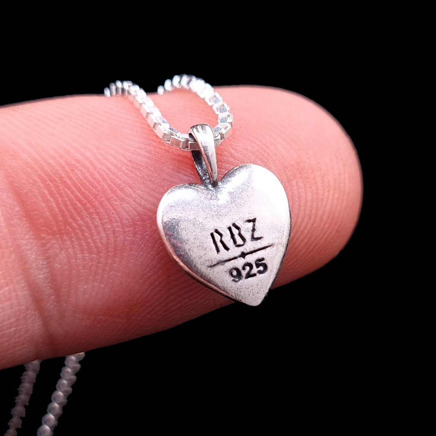 A close-up view of the back of the Sterling silver Gothic Ribcage Heart pendant from RBZ Jewelry being held on a finger for scale. The polished heart surface displays a clear engraving of the RBZ logo and the 925 sterling silver hallmark, attached to a delicate silver box chain.