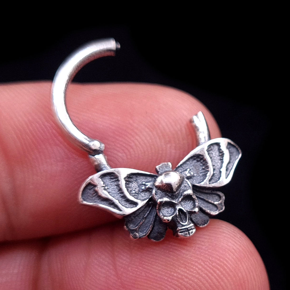 Sterling silver Gothic Death Moth septum ring shown in the open position, held between fingers to demonstrate the hinged clicker mechanism and the scale of the intricate skull and wing details, made by RBZ Jewelry.