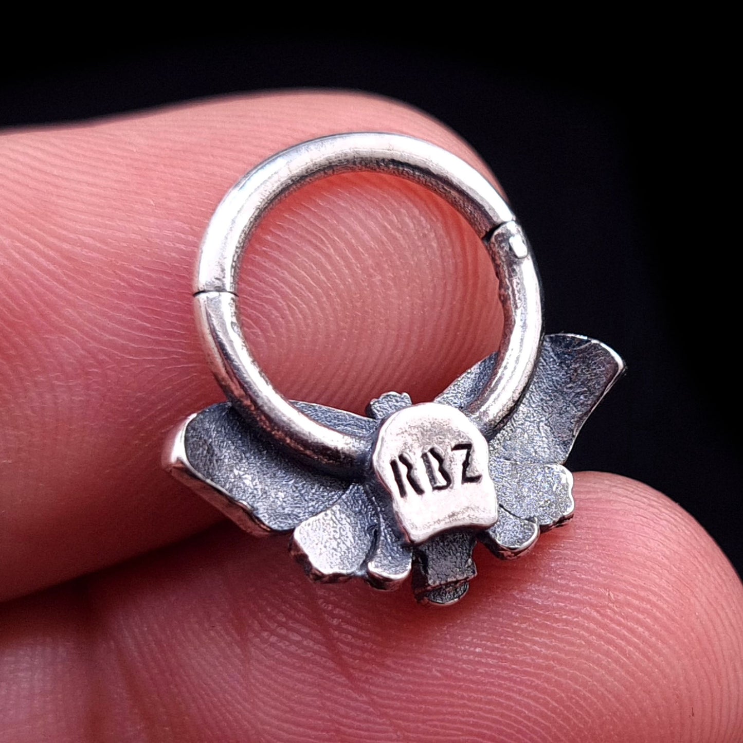 A sterling silver Death Moth septum clicker held between fingers, showcasing the back view with the RBZ brand hallmark and the textured finish of the wings.