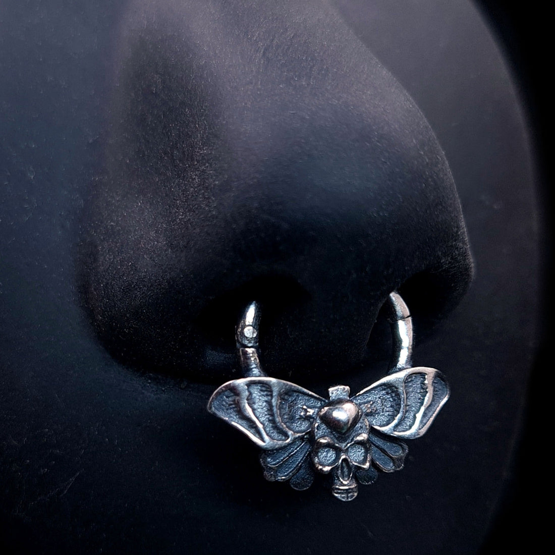 Sterling silver Gothic Death Moth septum clicker, by RBZ Jewelry,  worn on a black life-size silicone nose model, showcasing the symmetrical spread wings and skull motif in a realistic piercing placement.