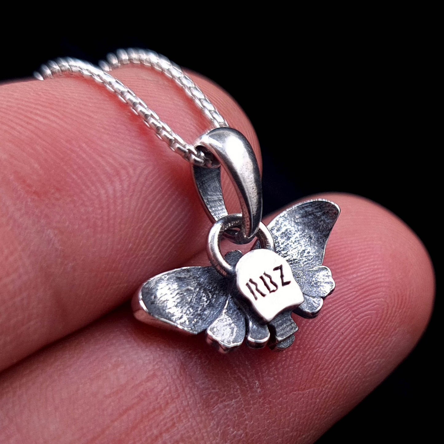 Back view of sterling silver Death Moth pendant held between fingers showing RBZ Jewelry logo engraving
