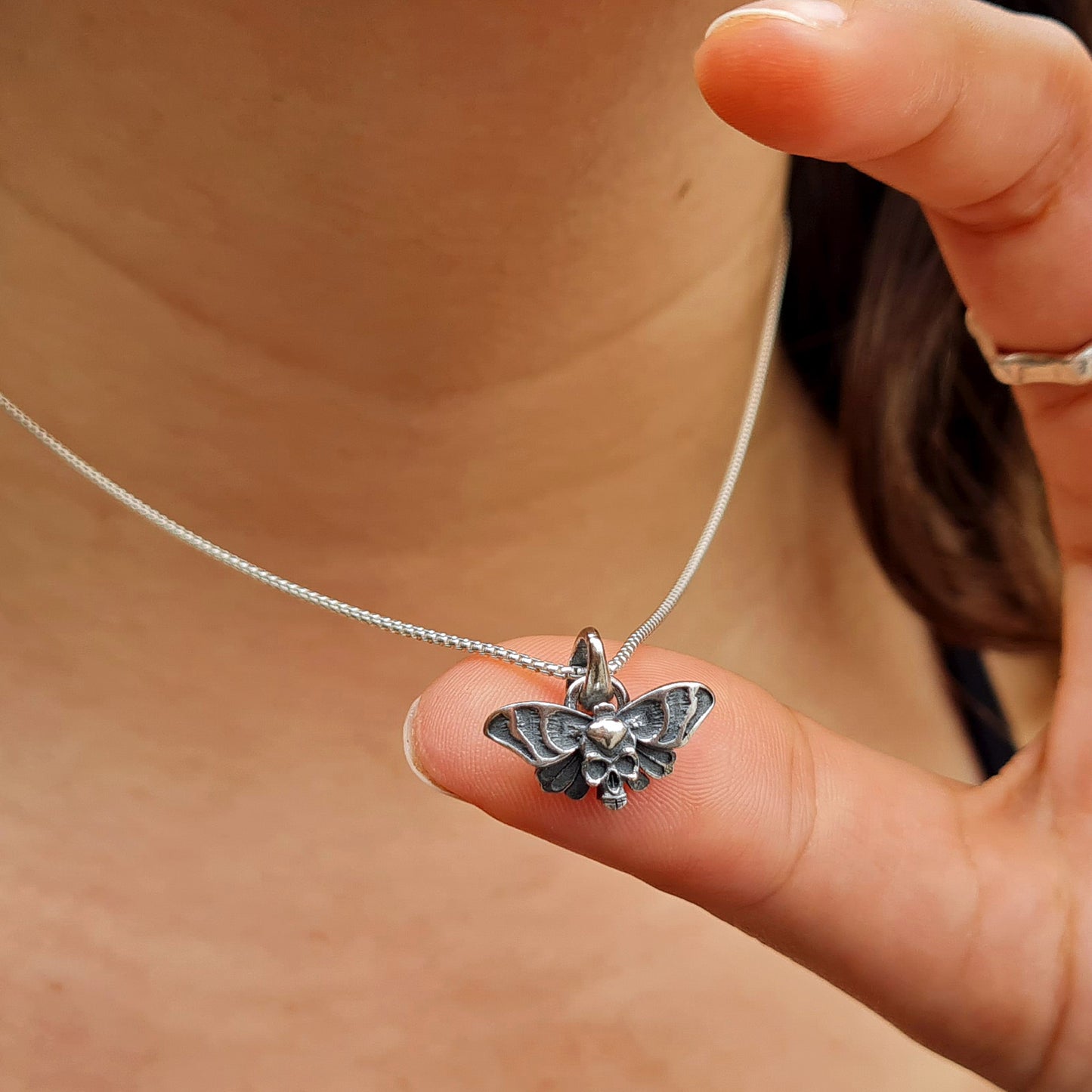 Sterling silver Death’s-head hawkmoth necklace held by a finger to demonstrate its small, delicate size and intricate details