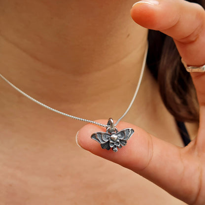 Sterling silver Death’s-head hawkmoth necklace held by a finger to demonstrate its small, delicate size and intricate details