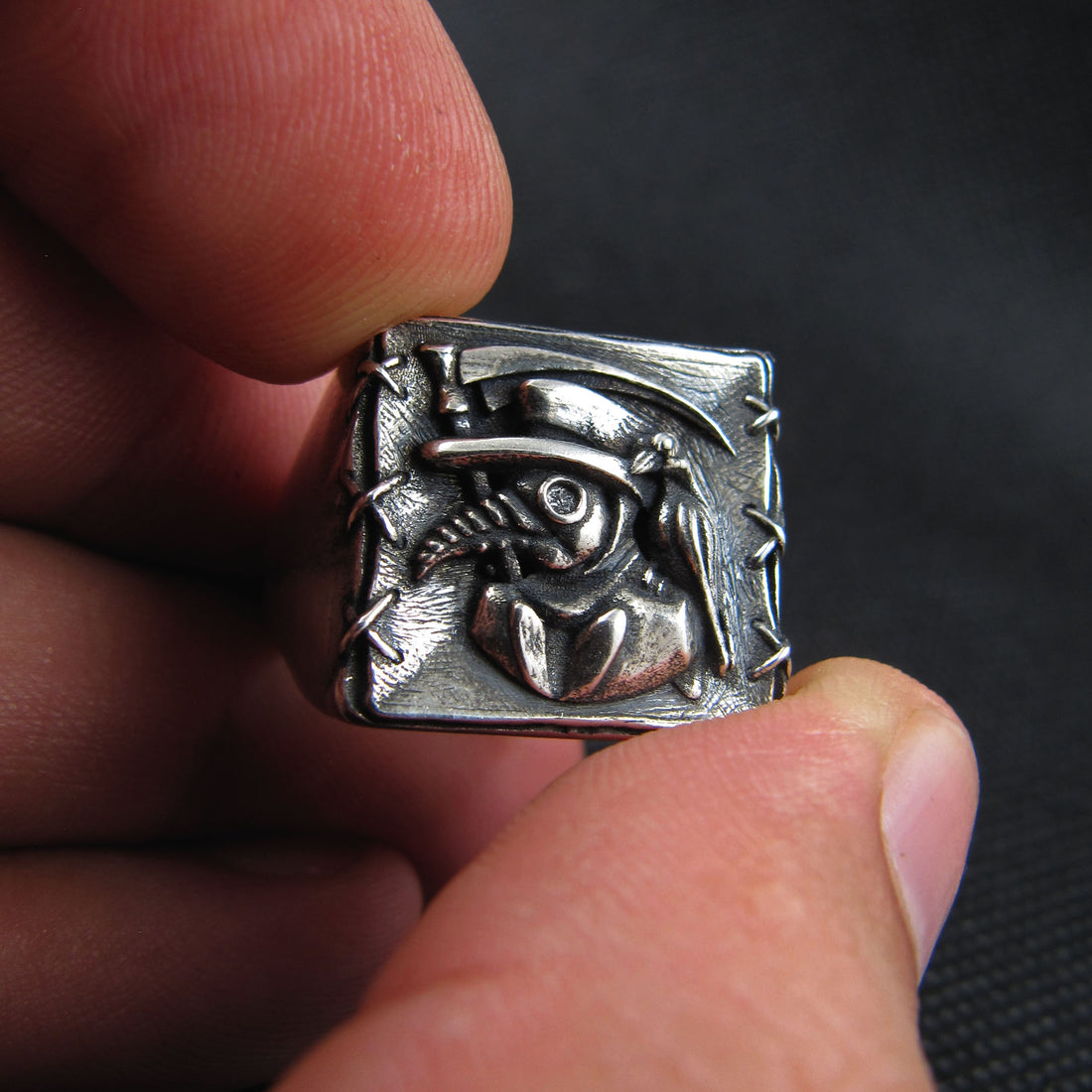 Sterling silver Plague Doctor signet ring by RBZ Jewelry held between two fingers to showcase the intricate scale and high-relief details of the beaked mask, raven, and sickle design.