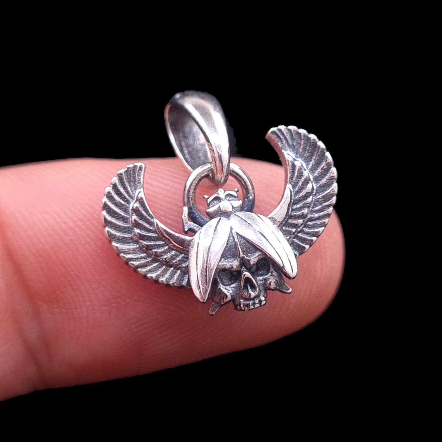 Front view of the sterling silver Gothic Egyptian Skull Scarab pendant by RBZ Jewelry, held on a finger for scale to show the size and intricate carving of the central skull and outstretched wings against a black background.