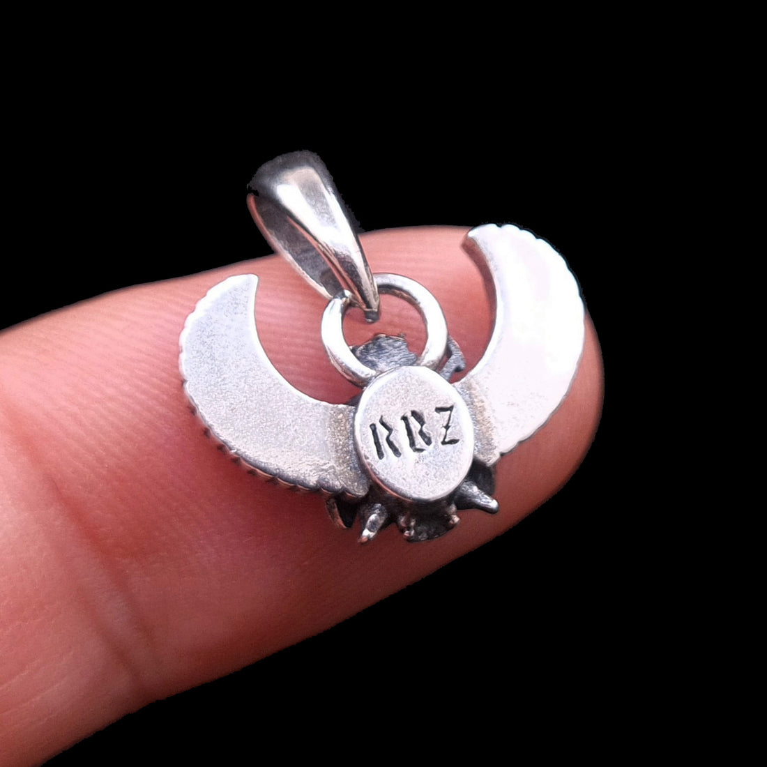 Rear view close-up of the sterling silver Gothic Egyptian Skull Scarab pendant from RBZ Jewelry, held on a finger for scale against a black background. The central circular medallion features the distinct, engraved stylized brand logo 'RBZ'.