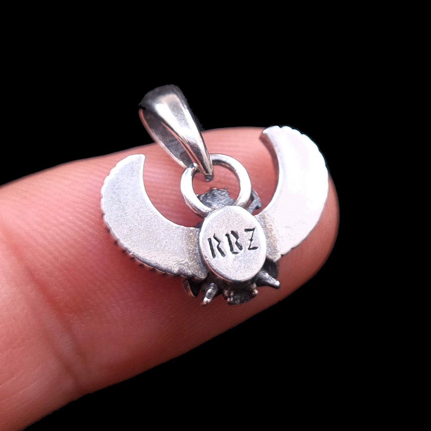 Rear view close-up of the sterling silver Gothic Egyptian Skull Scarab pendant from RBZ Jewelry, held on a finger for scale against a black background. The central circular medallion features the distinct, engraved stylized brand logo 'RBZ'.