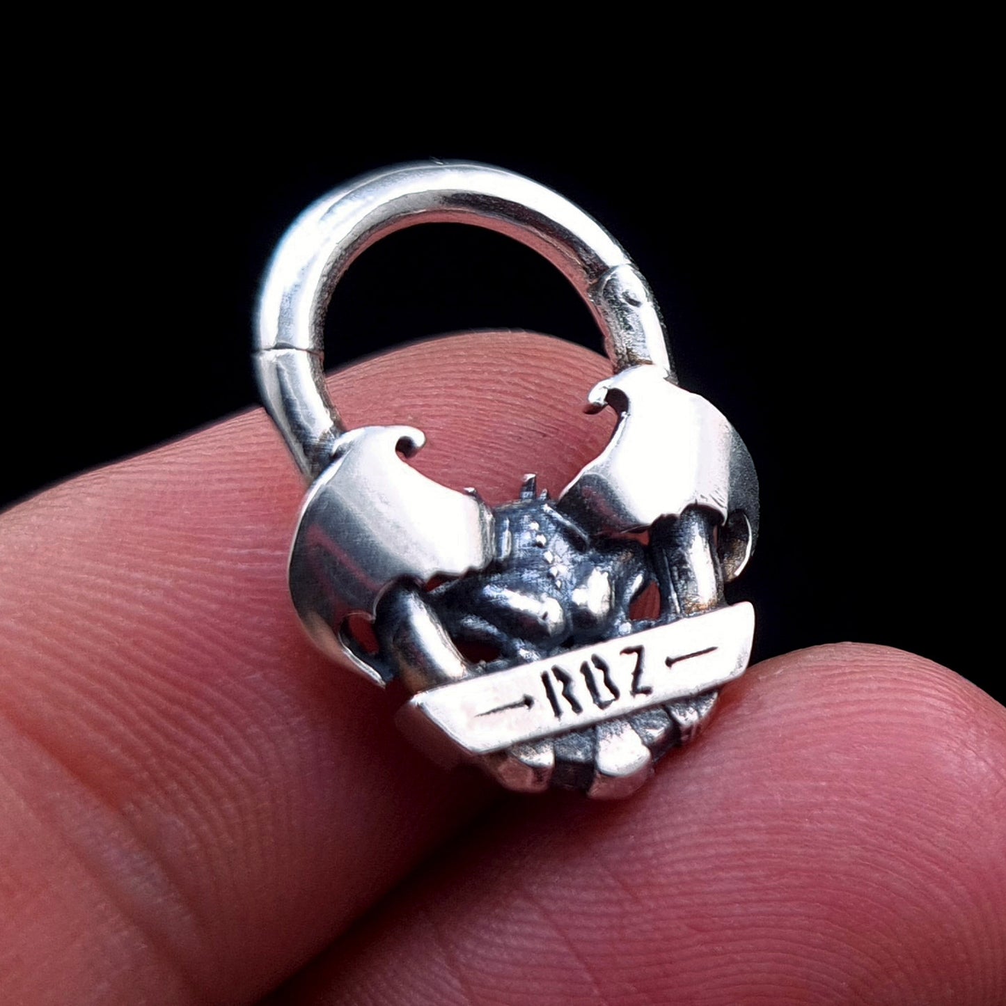 A back view of the RBZ Jewelry Gargoyle Guardian septum ring held between fingers, showing the RBZ logo engraved on the sterling silver banner and providing a sense of scale for the gothic winged design.