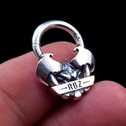 A back view of the RBZ Jewelry Gargoyle Guardian septum ring held between fingers, showing the RBZ logo engraved on the sterling silver banner and providing a sense of scale for the gothic winged design.