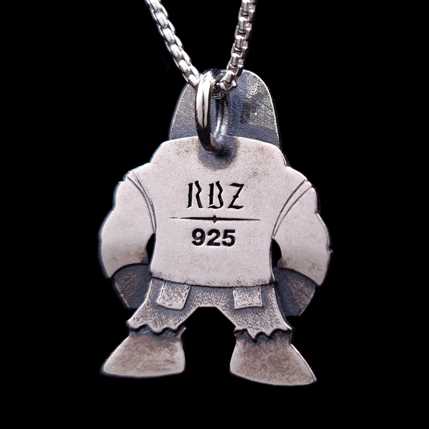 Back view of the RBZ Jewelry sterling silver Golem of Prague pendant, showing the flat, oxidized surface engraved with the "RBZ" logo and "925" silver hallmark. The silhouette mirrors the punk-rock guardian's form, suspended from a silver box chain against a black background.