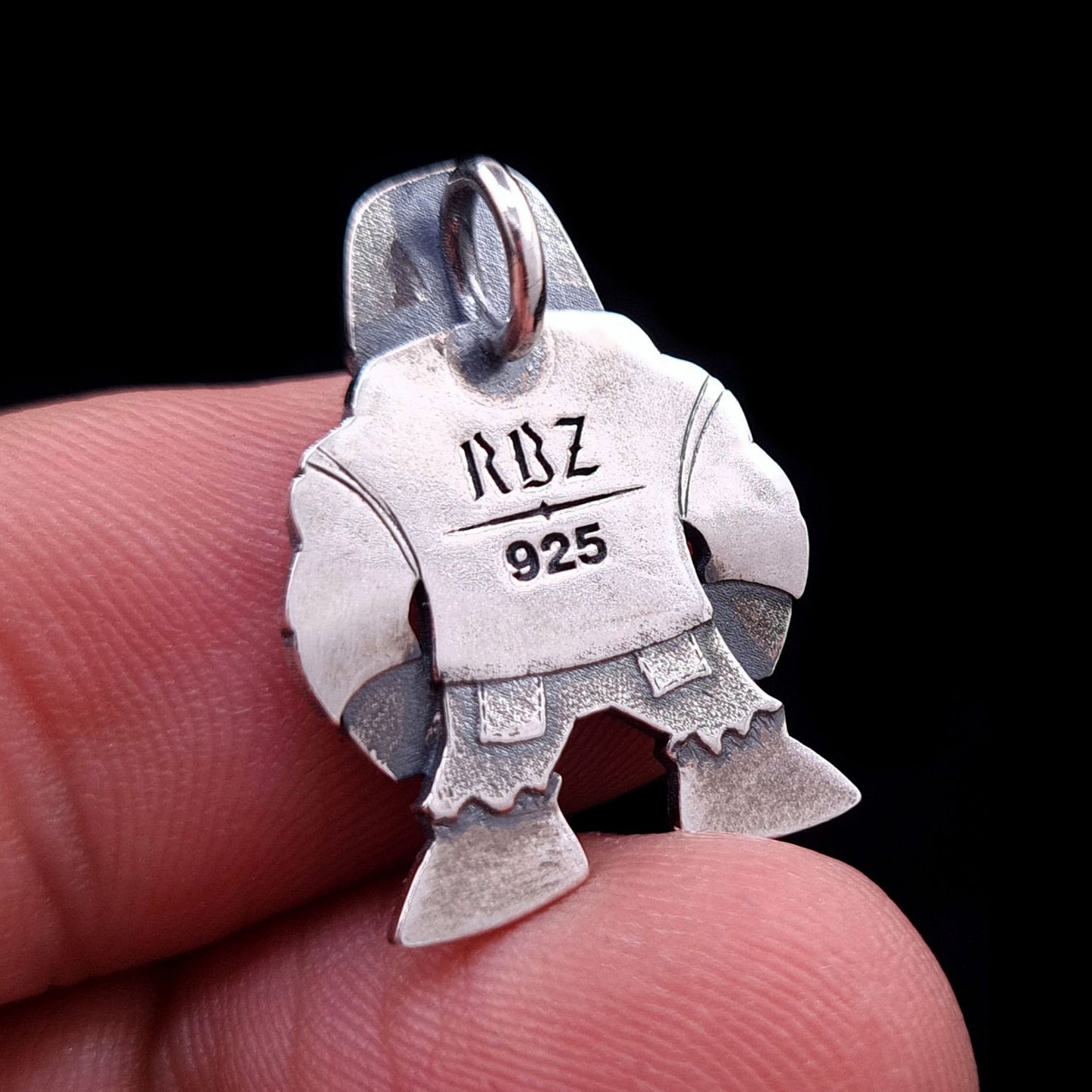 Back view of the handcrafted sterling silver Golem of Prague pendant by RBZ Jewelry, held on a finger for size reference. The oxidized silver surface features the engraved RBZ logo and 925 hallmark on the flat silhouette of the punk-rock guardian character.