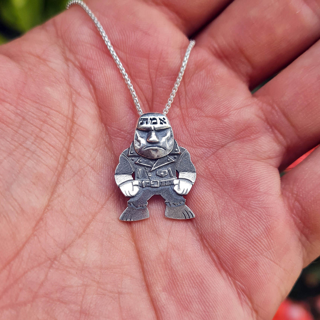 Handcrafted sterling silver Golem of Prague pendant by RBZ Jewelry, shown resting in a palm for size reference. The punk-rock guardian features a weathered leather jacket, clenched fists, and the Hebrew word אמת (Truth) on its forehead, attached to a silver chain.