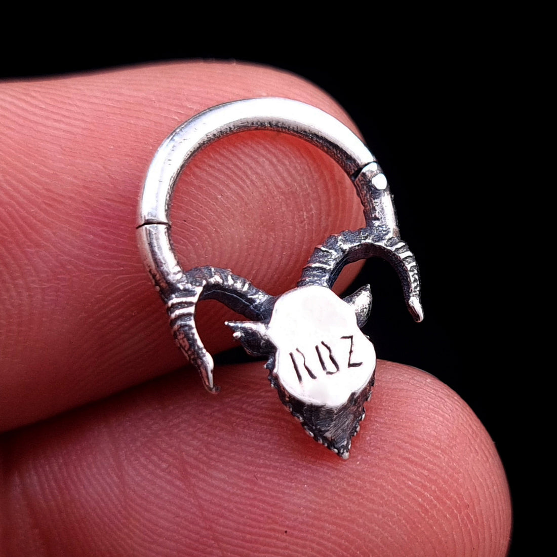Back view of the sterling silver Horned Demon septum ring by RBZ Jewelry, held between fingers in a closed position, showing the smooth backplate clearly engraved with "RBZ", the textured horns, and the ring loop against a black background.