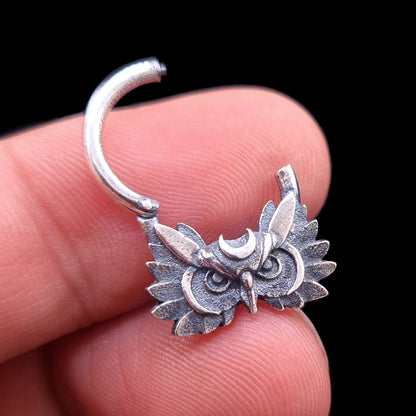 Handheld view of the Sterling silver Moon Owl septum ring by RBZ Jewelry, shown in the open position to demonstrate the clicker mechanism. The handcrafted owl face with a crescent moon and dark oxidized finish is held between fingers against a black background.