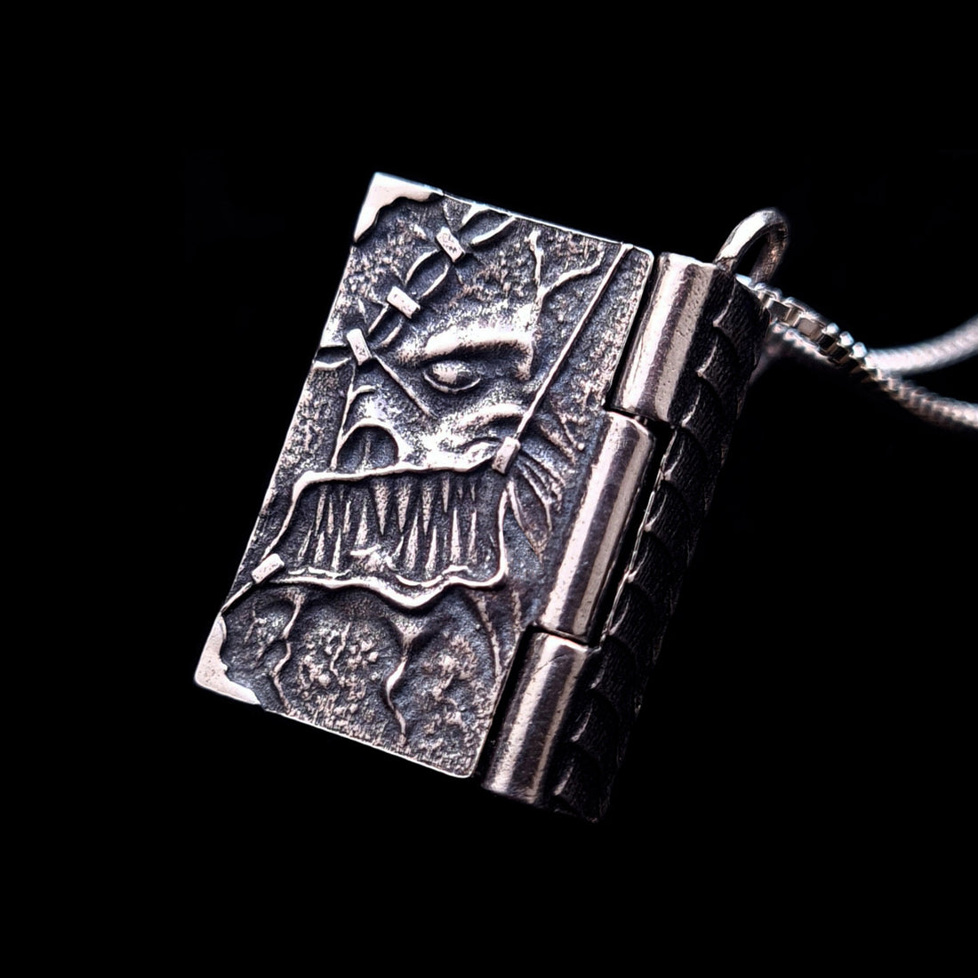 Detailed front view of a sterling silver Necronomicon book locket pendant against a black background. The oxidized silver cover features a high-relief "stitched skin" texture with a twisted skeletal face and jagged teeth. The design shows a functional heavy-duty hinge and a silver box chain, capturing the dark, gothic aesthetic of H.P. Lovecraft’s cosmic horror. Created by RBZ Jewelry.