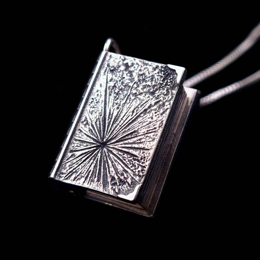 Back view of the sterling silver Necronomicon book locket pendant, featuring a deeply textured radial starburst or cosmic sigil design. The oxidized silver has a weathered, ancient finish, with the side of the pendant showing realistic page ridges. This gothic book-shaped locket is captured against a black background, highlighting the artisanal craftsmanship and Lovecraftian theme.