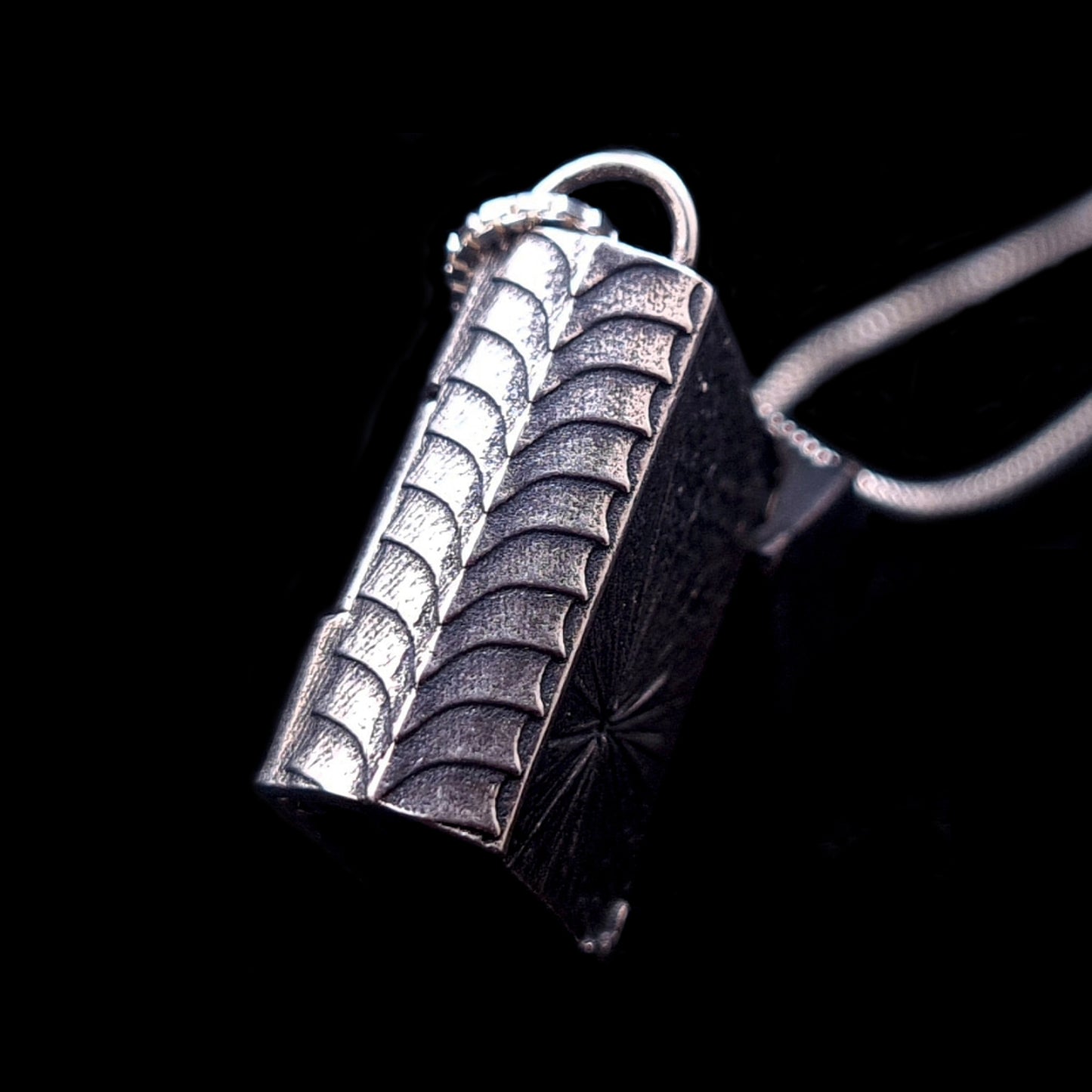 Side profile of the sterling silver Necronomicon book locket pendant, showcasing the intricately textured spine and heavy-duty hinge. The oxidized silver features a ribbed, scale-like pattern that evokes ancient, weathered bookbinding or visceral, reptilian skin. The image is set against a black background with a partial view of the silver box chain, highlighting the pendant's solid construction and macabre gothic aesthetic.
