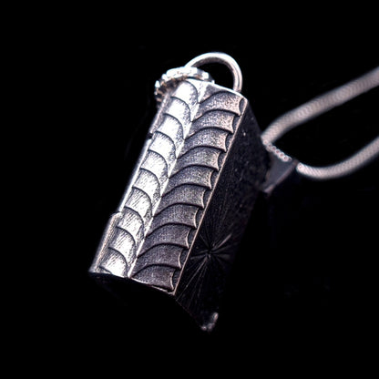 Side profile of the sterling silver Necronomicon book locket pendant, showcasing the intricately textured spine and heavy-duty hinge. The oxidized silver features a ribbed, scale-like pattern that evokes ancient, weathered bookbinding or visceral, reptilian skin. The image is set against a black background with a partial view of the silver box chain, highlighting the pendant's solid construction and macabre gothic aesthetic.