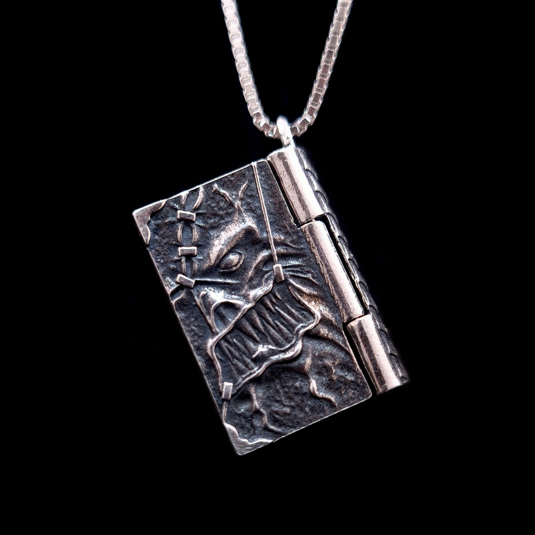 Front-angled view of the detailed sterling silver Necronomicon book locket pendant, shown hanging from a matching silver box chain. The cover features a visceral, high-relief skeletal face with jagged teeth and an intricate stitched skin texture in oxidized, weathered silver. The heavy-duty side hinge and the pendant's substantial side profile are prominent, showcasing its solid construction and gothic horror aesthetic against a black background.