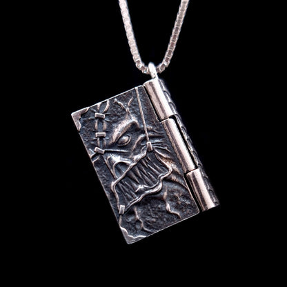 Front-angled view of the detailed sterling silver Necronomicon book locket pendant, shown hanging from a matching silver box chain. The cover features a visceral, high-relief skeletal face with jagged teeth and an intricate stitched skin texture in oxidized, weathered silver. The heavy-duty side hinge and the pendant's substantial side profile are prominent, showcasing its solid construction and gothic horror aesthetic against a black background.