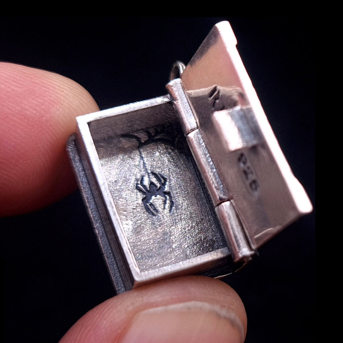A person’s fingers hold an open sterling silver Necronomicon book locket to show its small, pendant-sized proportions. The interior reveals a detailed secret spider engraving on the textured bottom of the hollow compartment. The polished inner lid shows the RBZ brand logo and 925 hallmark, while the side of the silver pendant features realistic book page ridges, emphasizing the artisanal, gothic horror design.