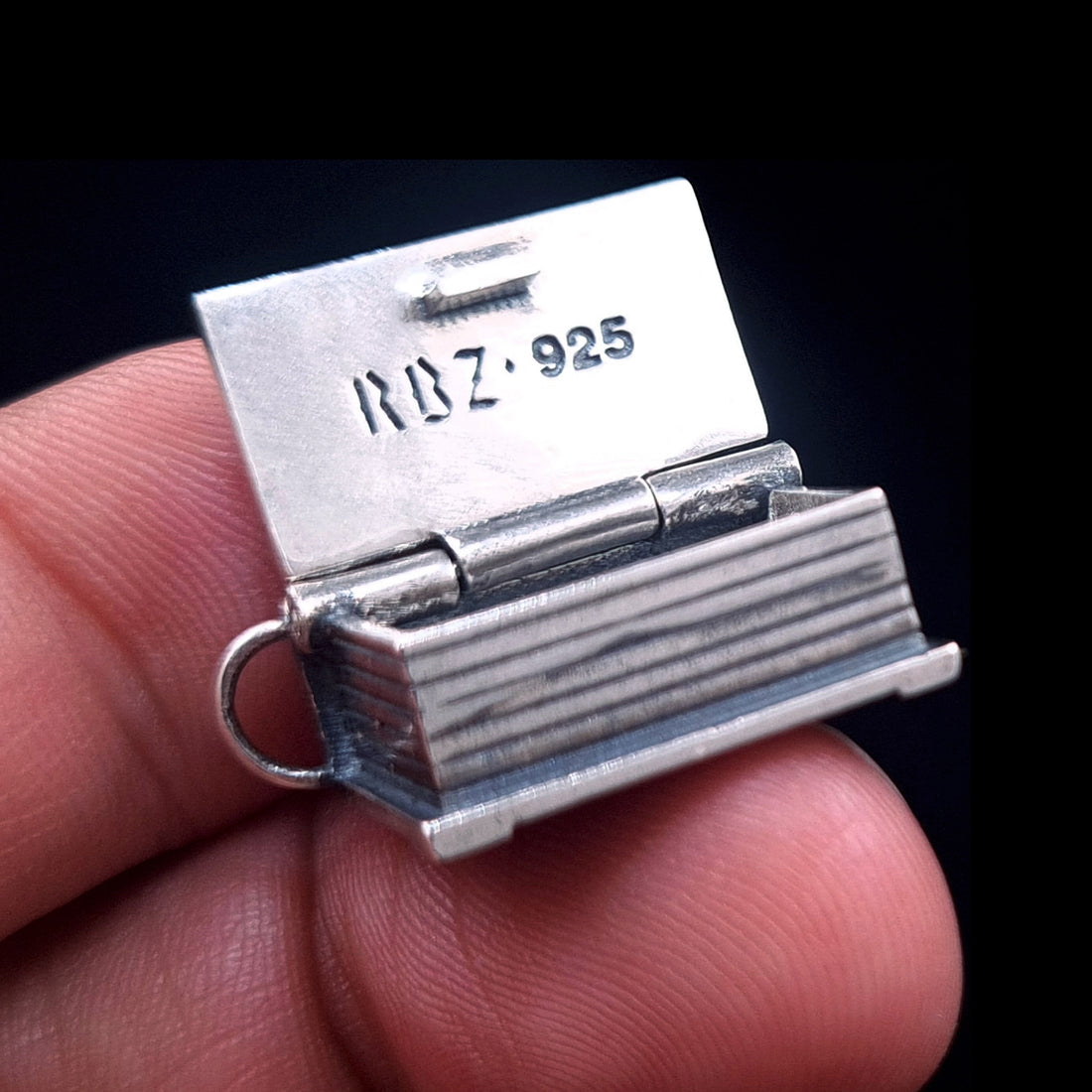 An open sterling silver Necronomicon book locket held between two fingers to demonstrate its scale and proportions. The polished inner lid clearly displays the RBZ brand logo and 925 hallmark engraving. This view highlights the realistic page ridges on the side of the pendant and the sturdy hinge construction, showcasing the artisanal gothic horror craftsmanship of RBZ Jewelry against a dark background.