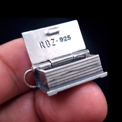 An open sterling silver Necronomicon book locket held between two fingers to demonstrate its scale and proportions. The polished inner lid clearly displays the RBZ brand logo and 925 hallmark engraving. This view highlights the realistic page ridges on the side of the pendant and the sturdy hinge construction, showcasing the artisanal gothic horror craftsmanship of RBZ Jewelry against a dark background.