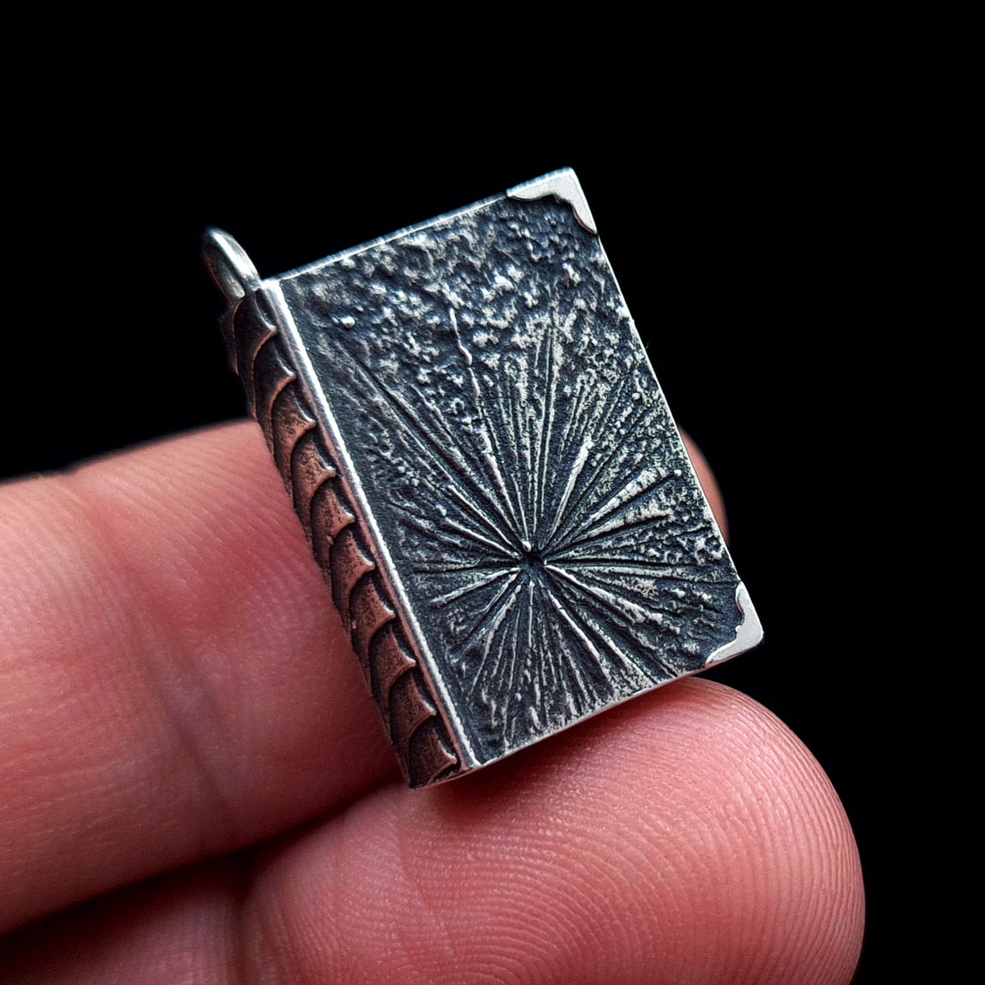 A person's fingers hold the back of the sterling silver Necronomicon book locket to demonstrate its compact, pendant-sized scale. The oxidized silver surface features a deeply textured, ancient-looking cosmic sigil or starburst design. This view highlights the contrast between the weathered relief and the detailed scale-like spine, emphasizing the artisanal, dark fantasy quality of the RBZ Jewelry piece.