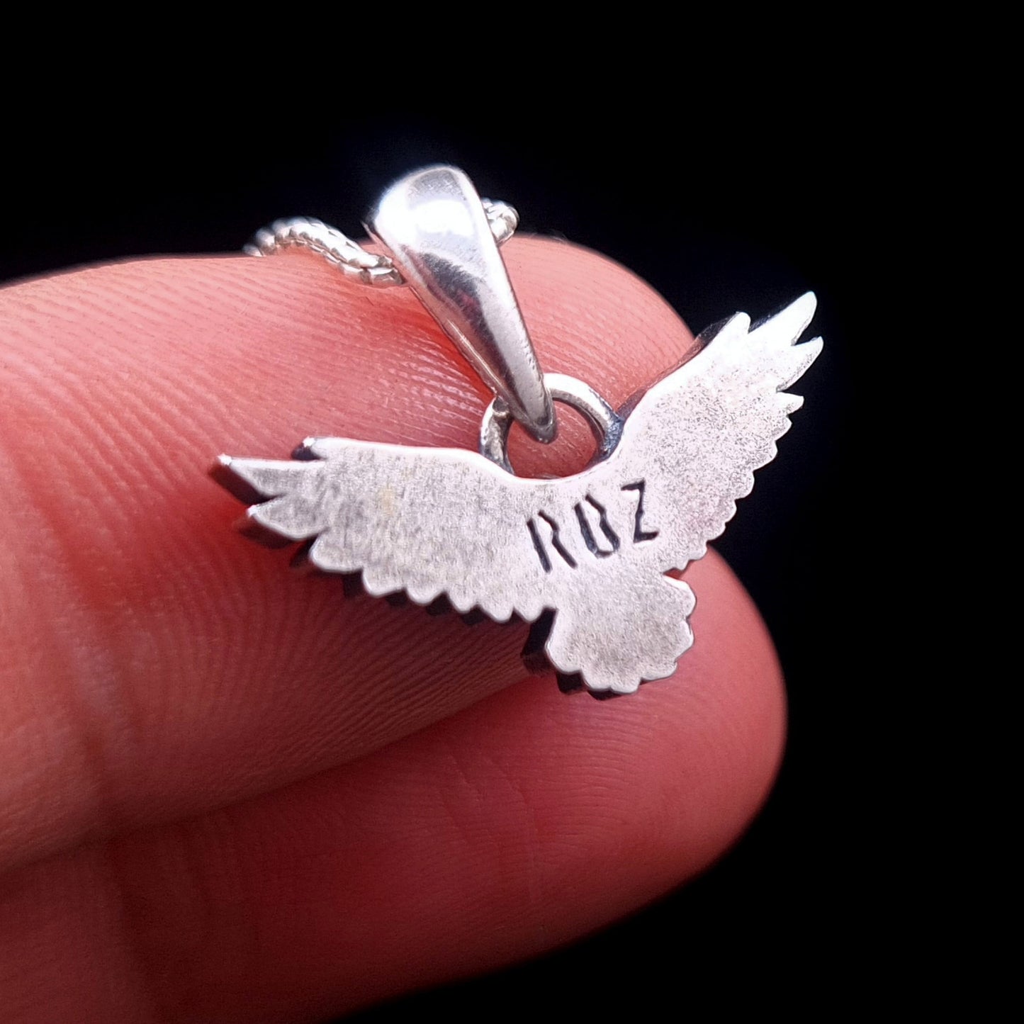 Back view of the sterling silver flying raven pendant held on fingers for scale, showing the smooth reverse side with RBZ brand engraving, handcrafted Gothic crow necklace by RBZ Jewelry.