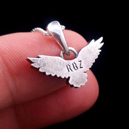 Back view of the sterling silver flying raven pendant held on fingers for scale, showing the smooth reverse side with RBZ brand engraving, handcrafted Gothic crow necklace by RBZ Jewelry.