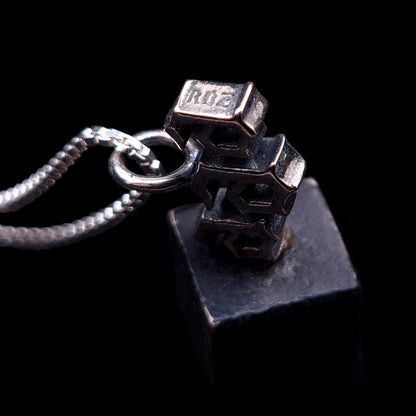 Detailed macro photograph, from a side angle, of the Sterling silver Gothic Satanic 666 Number of the Beast pendant from RBZ Jewelry. The image shows the thickness and dark, oxidized patina on the edges of the blocky, gothic-font numbers. A sharp, crisp engraving of the logo text 'RBZ' is clearly visible on a side surface of the first number '6'. The pendant is attached to a silver box chain and rests on a dark, rough metal prop against a solid black background, showcasing the handcrafted detail.
