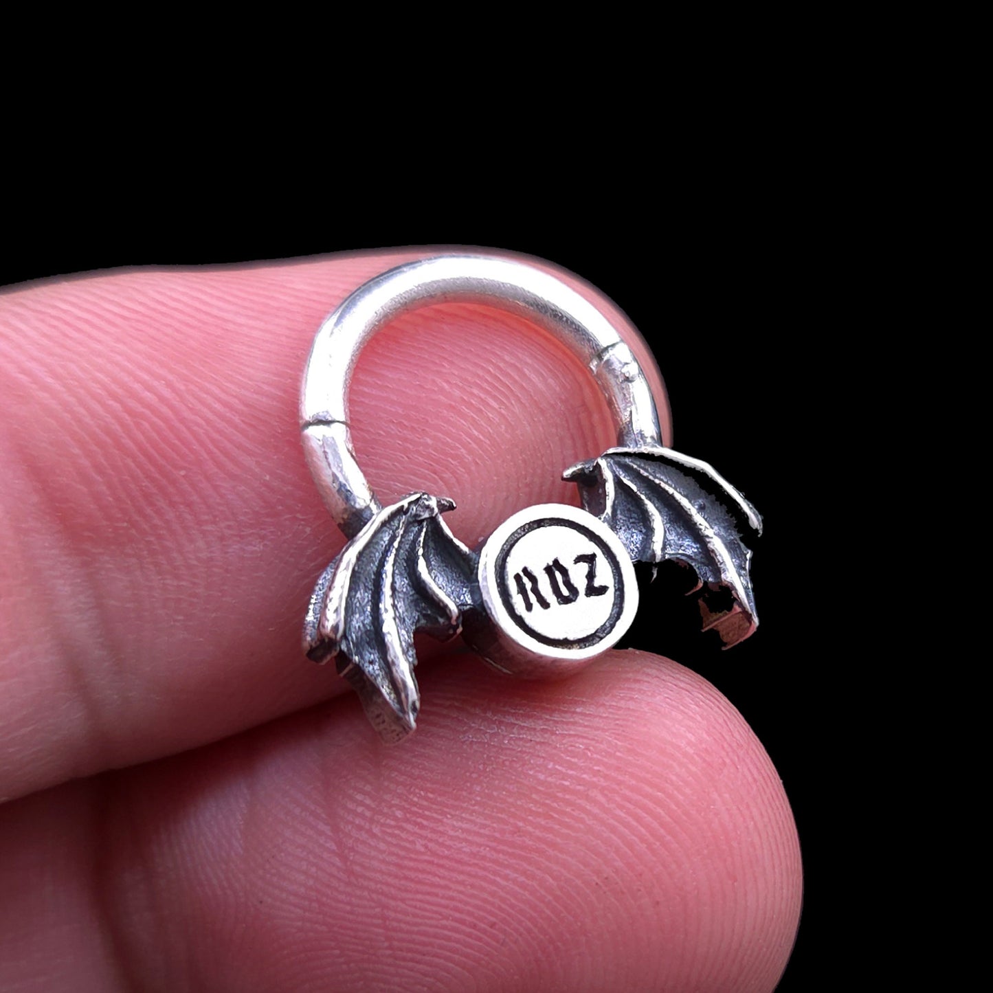 Rear view of the gothic 925 sterling silver winged septum clicker ring by RBZ Jewelry, held open between two fingers against a black background to show the engraved RBZ brand logo, scale, and size proportions.