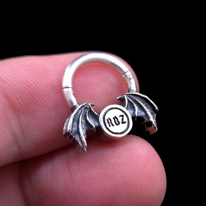 Rear view of the gothic 925 sterling silver winged septum clicker ring by RBZ Jewelry, held open between two fingers against a black background to show the engraved RBZ brand logo, scale, and size proportions.